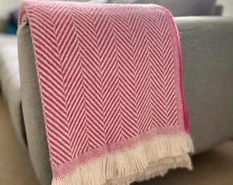 Puede incluir: Una manta de lana de color rosa y blanco con un patrón de espiga y un borde de flecos. La manta está colocada sobre un sofá gris.