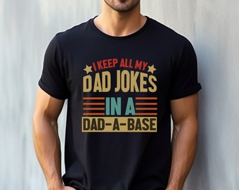 Op de afbeelding: Een zwart T-shirt met een retro-stijl grafische afbeelding met de tekst "I keep all my dad jokes in a dad-a-base".