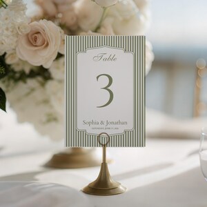 May include: A table number card with a green and white striped background. The card features the number 3 in a large font, with the names "Sophia & Jonathan" and the date below. The card is held by a gold-colored metal stand.