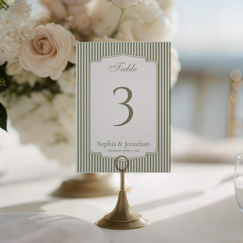 May include: A table number card with a green and white striped background. The card features the number 3 in a large font, with the names "Sophia & Jonathan" and the date below. The card is held by a gold-colored metal stand.