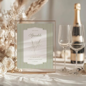 May include: A framed invitation with the text "Let Love Sparkle" and the names Sophia & Jonathan. The invitation is set on a table with champagne flutes and a champagne bottle. The frame is gold.