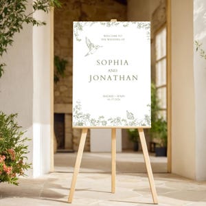 May include: A wooden easel displays a wedding welcome sign. The sign is white with green floral and hummingbird illustrations. Text reads "Welcome to the Wedding of Sophia and Jonathan" with the date and location, Madrid, Spain, 06-17-2026.