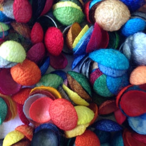 Könnte beinhalten: Nahaufnahme einer Ansammlung farbenfroher Filzformen aus Wolle. Das Bild zeigt eine Vielzahl von Farben, darunter Rot, Orange, Gelb, Grün, Blau und Lila. Die Filzstücke sind kleine, abgerundete Formen, einige mit geschichteten Designs, was eine strukturierte Komposition ergibt.