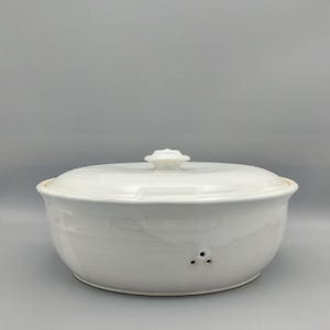 Könnte beinhalten: Eine weiße Keramik-Auflaufform mit Deckel. Die Form hat eine runde Form mit leicht ausgestelltem Rand und einem Deckel mit dekorativem Knauf. Drei kleine Löcher sind an der Seite der Form sichtbar.