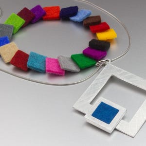 May include: A modern necklace featuring a silver wire chain and a geometric pendant. The pendant has a square frame with a smaller square inset with blue fabric. The necklace also includes a colorful array of felt squares in various colors.