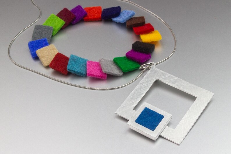 May include: A modern necklace featuring a silver wire chain and a geometric pendant. The pendant has a square frame with a smaller square inset with blue fabric. The necklace also includes a colorful array of felt squares in various colors.