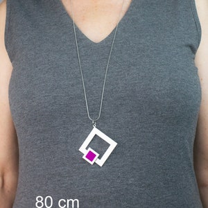 May include: A silver necklace with a geometric pendant. The pendant features a white square frame with a smaller magenta square inside. The necklace is 80 cm long.