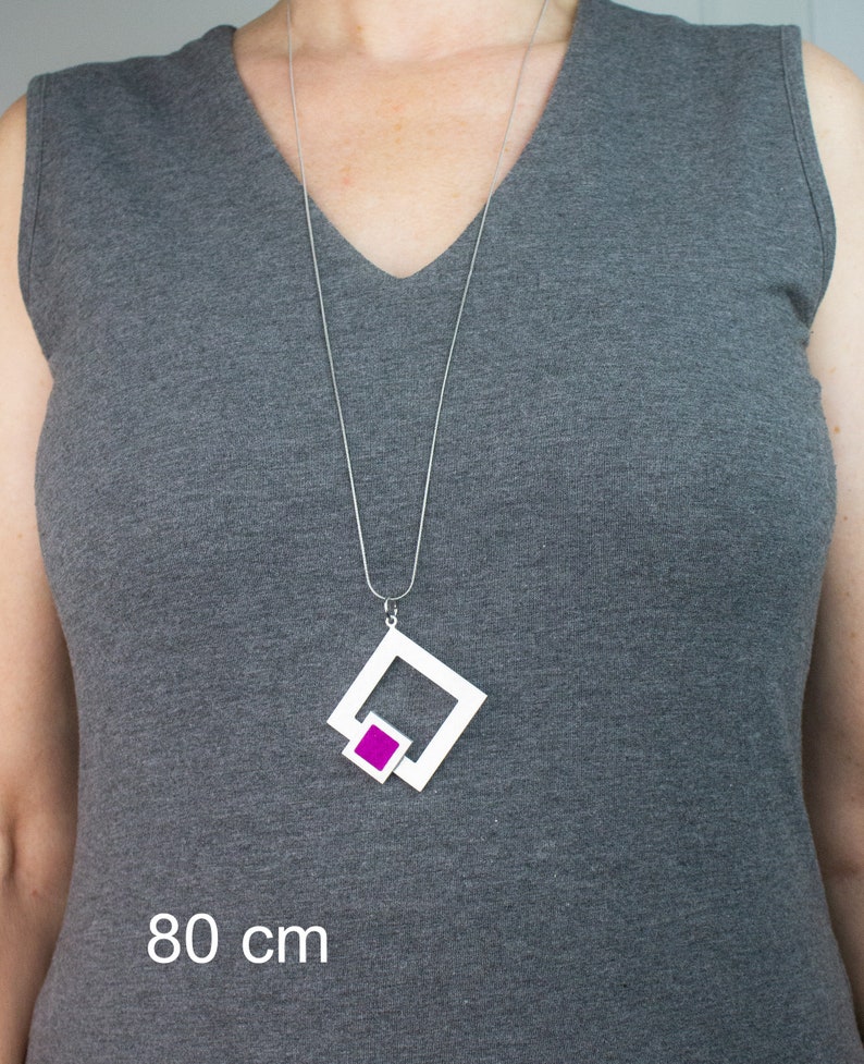 May include: A silver necklace with a geometric pendant. The pendant features a white square frame with a smaller magenta square inside. The necklace is 80 cm long.