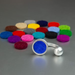 Könnte beinhalten: Ein silberner Ring mit einem blauen Filzkreis und einem kleinen silbernen Ball. Um den Ring herum sind mehrere bunte Filzkreise in verschiedenen Farben, darunter Rot, Rosa und Grün, verstreut. Der Hintergrund ist einfarbig grau.
