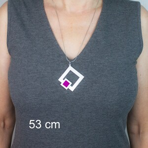 May include: A silver necklace with a white geometric pendant featuring a magenta square. The necklace is displayed on a person wearing a gray sleeveless top. The image includes the text "53 cm".
