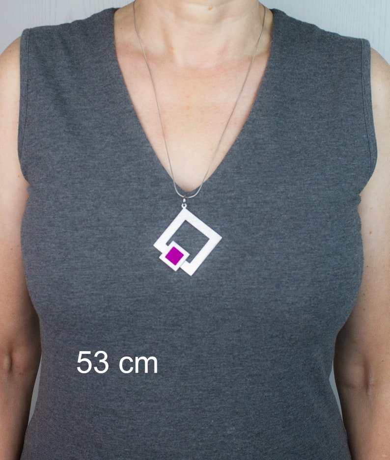 May include: A silver necklace with a white geometric pendant featuring a magenta square. The necklace is displayed on a person wearing a gray sleeveless top. The image includes the text "53 cm".