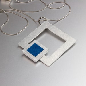 May include: A silver-toned pendant necklace featuring a geometric design. The pendant has a large square frame with a smaller square inset, filled with blue material. The necklace chain is a thin, silver snake chain.