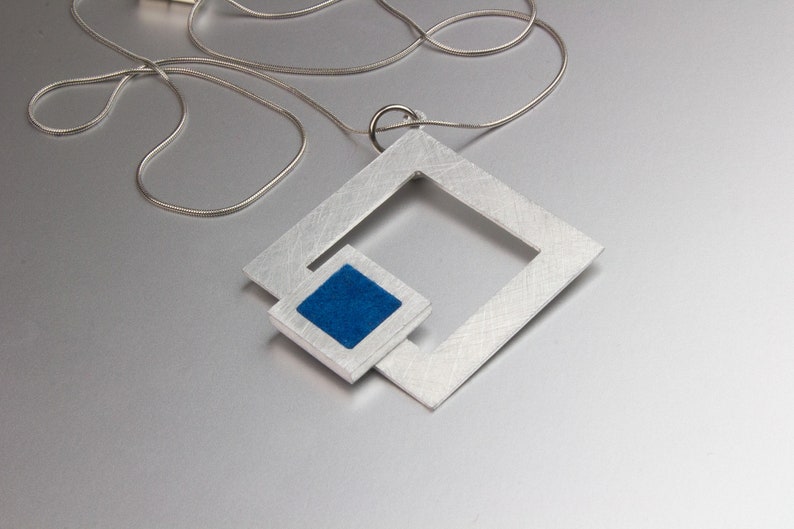 May include: A silver-toned pendant necklace featuring a geometric design. The pendant has a large square frame with a smaller square inset, filled with blue material. The necklace chain is a thin, silver snake chain.