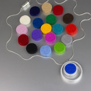 Könnte beinhalten: Eine silberne Halskette mit einem Anhänger, der einen blauen Filzkreis in einem silbernen Ring zeigt. Die Halskette ist von einer Auswahl farbiger Filzkreise in verschiedenen Farben umgeben, darunter Rot, Grün und Gelb. Die Halskette liegt auf einer silbernen Oberfläche.