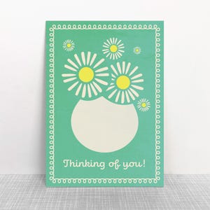 Könnte beinhalten: Ein rechteckiges, türkisfarbenes Holzschild mit weißem Rand und dem Schriftzug "Thinking of you!" in Schreibschrift. Das Schild zeigt eine weiße Vase mit Gänseblümchen und gelben Zentren.