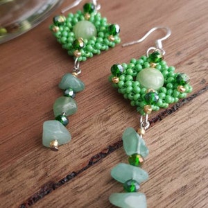 May include: A pair of green beaded earrings with a green stone pendant. The earrings are made with small green beads and a larger green stone in the centre. The earrings are hanging from silver hooks.