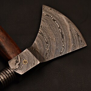 May include: A hand-forged tomahawk with a wooden handle and a damascus steel head. The head has a distinctive wavy pattern.