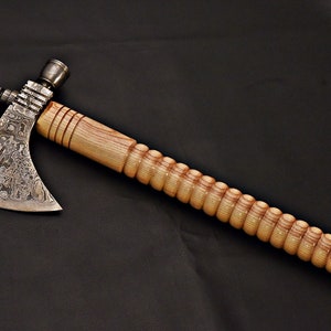 Puede incluir: Un hacha de guerra con un mango de madera y una hoja de acero con patrón. El mango es de madera marrón claro con una superficie texturizada. La hoja es de acero con un patrón de remolinos y líneas.