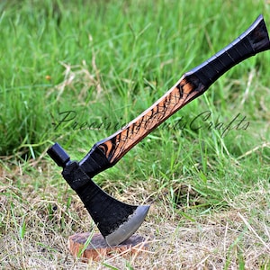 May include: A black and brown tomahawk with a wooden handle. The handle has a black and brown wood grain pattern. The tomahawk is sitting on a bed of green grass.
