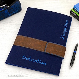 Könnte beinhalten: Ein marineblaues Filzportfolio mit einem braunen Lederriemen. Der Name "Sebastian" ist blau gestickt. Das Wort "Zeugnisse" ist ebenfalls blau gestickt. Ein schwarzer Stift befindet sich auf der rechten Seite.