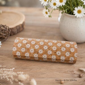 Könnte beinhalten: Ein zylindrisches Etui aus Kork mit einem sich wiederholenden Muster aus kleinen weißen Gänseblümchen. Die Außenfläche hat eine natürliche, hellbraune Korkstruktur mit einem durchgehenden Blumenmuster. Der Text Handmade for Killefitz ist sichtbar.
