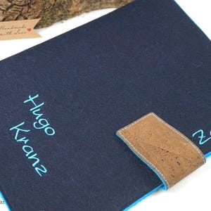 Könnte beinhalten: Eine marineblaue Filz-Notizbuchhülle mit dem Namen "Hugo Kranz" in Türkis gestickt. Die Hülle hat einen braunen Korkverschluss und einen türkisfarbenen Rand. Das Wort "Zeugnisse" ist ebenfalls in Türkis gestickt.