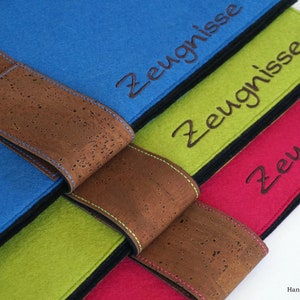 Könnte beinhalten: Vier Filzordner mit Korkbändern, jeder Ordner ist in einer anderen Farbe: blau, grün, rot und pink. Die Ordner sind übereinander gestapelt. Jeder Ordner hat das Wort "Zeugnisse" in schwarzem Faden auf der Vorderseite gestickt.