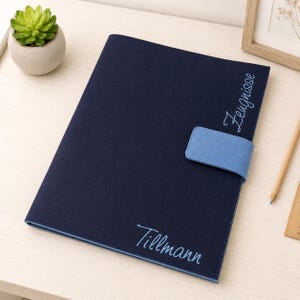 Könnte beinhalten: Eine dunkelblaue Dokumentenmappe aus Filz mit einem hellblauen Verschlussriemen. Die Mappe ist mit dem Text "Zeugnisse" in Schreibschrift oben rechts und dem Namen "Tillmann" in derselben Schriftart unten mittig bestickt.