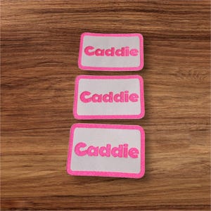 Op de afbeelding: Drie witte rechthoekige plekken met roze randen en het woord "Caddie" in roze letters.