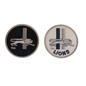 May include: Two embroidered patches with a lion design. The patches are circular and have a black and white background. The lion is white and the text "TM LIONS" is in blue.