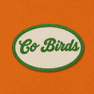 Può includere: Una toppa ovale bianca con un bordo verde e il testo "Go Birds" in lettere corsive verdi.