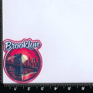 May include: A red and white embroidered patch with the word "Brooklyn" in a white script font. The patch features a depiction of the Brooklyn Bridge against a sunset backdrop.
