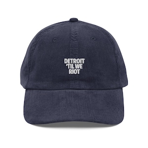 May include: Navy blue corduroy baseball cap with the text "DETROIT 'TIL WE RIOT" embroidered in white. The cap has a curved brim and a rounded crown. A casual accessory for everyday wear.