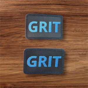 Peut inclure: Deux patchs en tissu noir avec des coutures blanches autour des bords. Les patchs ont le mot "GRIT" brodé en fil bleu.