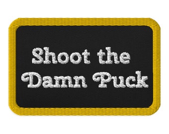 May include: Embroidered patch with the text "Shoot the Damn Puck" in white script on a black background. The patch has a yellow border and is rectangular with rounded corners.