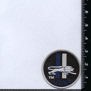 Op de afbeelding: Een geborduurde patch in zwart en wit met een leeuw en het woord "LIONS" in een witte cirkel. De leeuw rent naar rechts en heeft een blauwe en witte streep achter zich. Het woord "TM" staat boven de leeuw.