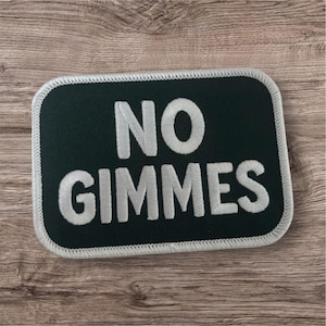 May include: A rectangular patch with rounded corners, featuring the words "NO GIMMES" in white embroidered letters on a dark green background. The patch has a white border and is set against a wood grain surface.