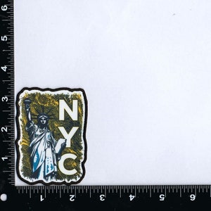 May include: A black and yellow patch with a white outline featuring a blue and white illustration of the Statue of Liberty and the text "NYC".