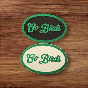 Puede incluir: Dos parches bordados de forma ovalada con costuras verdes. Ambos parches tienen el texto "Go Birds" en letras cursivas verdes. Un parche tiene un fondo negro y el otro un fondo blanco.