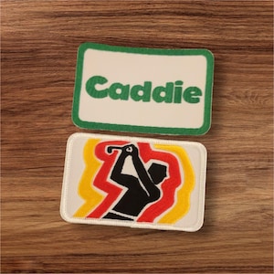 May include: Two embroidered patches on a wooden surface. The top patch has the word "Caddie" in green on a white background with a green border. The bottom patch features a golfer in black, with red and yellow accents.