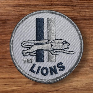 May include: A round, embroidered patch featuring a running lion in silver thread. The patch has a light gray background with two vertical stripes, one navy blue and one silver. The word "LIONS" is embroidered in navy blue.