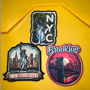 May include: Three embroidered patches with New York City themes. The first patch is black with a white outline and features the Statue of Liberty and the text "New York City". The second patch is red with a white outline and features the Brooklyn Bridge and the text "Brooklyn". The third patch is black with a white outline and features the Statue of Liberty and the text "NYC".