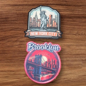 May include: Two embroidered patches featuring New York City landmarks. The top patch is a blue and white patch with the Statue of Liberty and the text "NEW YORK CITY". The bottom patch is a red and white patch with the Brooklyn Bridge and the text "Brooklyn".