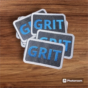 Könnte beinhalten: Ein Stapel aus fünf grauen Stoff-Patches mit weißem Rand. Jeder Patch hat das Wort "GRIT" in Blau gestickt.