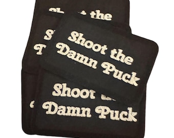 May include: A pile of black rectangular patches with the words "Shoot the Damn Puck" embroidered in white script. The patches are stacked on a white background.