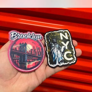 May include: Two embroidered patches, one pink with a sunset view of the Brooklyn Bridge and the word "Brooklyn" in white, and the other black with a yellow outline and the Statue of Liberty and the letters "NYC" in white.
