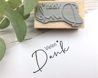 May include: A wooden stamp with a gray rubber surface that reads "Vielen Dank" with a small heart symbol. The stamp is used to create a black ink impression on a white card that reads "Vielen Dank" with a small heart symbol.