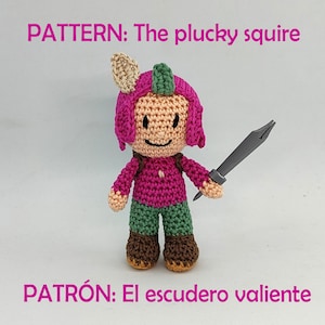 May include: A crocheted amigurumi toy of a pink-haired character with a green hat, brown boots, and a gray sword. The toy is holding the sword in its right hand. The text "PATTERN: The plucky squire" is above the toy and "PATRÓN: El escudero valiente" is below the toy.