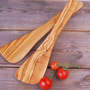 Può includere: Due spatole di legno di ulivo, con un motivo a venature del legno naturale, sono appoggiate su una superficie di legno. Tre pomodori rossi sono sulla superficie accanto alle spatole.