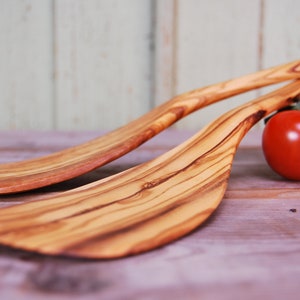 Könnte beinhalten: Zwei Holzspatel aus Olivenholz mit natürlicher Holzmaserung. Die Spatel liegen auf einer Holzoberfläche mit drei roten Tomaten im Hintergrund.